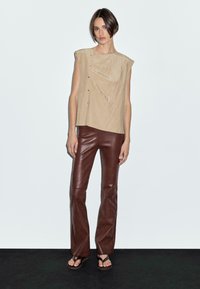 Beige ärmelloses Top mit asymmetrischem Saum und Knopfdetails, kombiniert mit engen braunen Lederhosen. Glatte Texturen und maßgeschneiderte Passform.