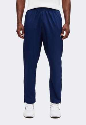 Personne portant un pantalon de jogging bleu marine et des baskets blanches, debout devant un fond clair uni.