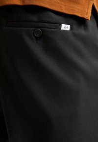 Svarta byxor med en framficka, en enkel knappstängning och en liten vit etikett med svart text. Tygets struktur är slät.