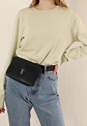 Borsa a tracolla nera in pelle con patta e hardware argentato, indossata sopra un maglione chiaro e jeans blu a vita alta.