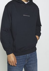 Námořnický pulovr s kapucí, přední kapsou a bílým logem "MENNACE". Vyrobeno z měkké směsi bavlny.