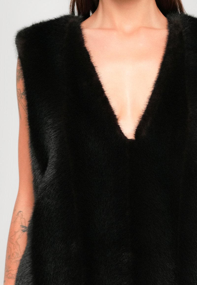 Sleeveless black fur vest with deep V neckline worn by tattooed person showing upper chest and arms.