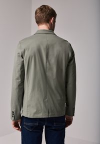 Chaqueta ligera de color verde oliva con un ajuste entallado, mangas largas, cuello con muesca y puños abotonados, combinada con jeans de mezclilla oscura.