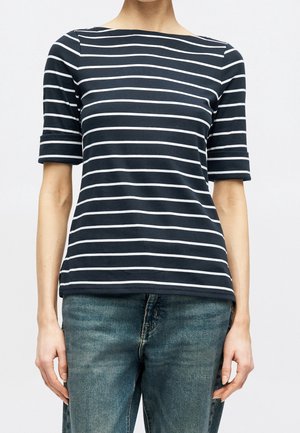 Person iført en mørkeblå og hvid vandretstribet kortærmet skjorte kombineret med falmede blå jeans, stående mod en ensfarvet baggrund.