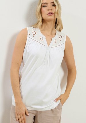 Donna con canotta bianca senza maniche con dettagli a occhiello floreali vicino alle spalle, pantaloni beige, in piedi contro un muro semplice.