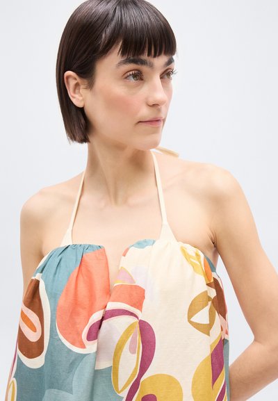 Γυναίκα με κοντά σκούρα μαλλιά, που φοράει τοπ με λαιμόκοψη halter και πολύχρωμα αφηρημένα σχέδια, κοιτάζοντας ελαφρώς προς τα δεξιά σε απλό φόντο.