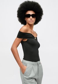 Kobieta o fryzurze afro, ubrana w czarną bluzkę bez ramiączek, szare spodnie w prążki i czarne okulary przeciwsłoneczne, stojąca z rękami w kieszeniach.