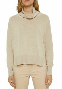 Kvinna klädd i en beige långärmad lös tröja med en bred krage och matchande beiga byxor, stående med armarna avslappnat vid sidorna.