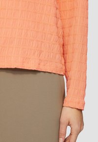 Pfirsichfarbener, strukturierter Langarmoberteil mit horizontalen Nähten und gerafftem Saum, kombiniert mit einer eng anliegenden, neutralen Hose.