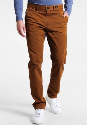 Mężczyzna w brązowych spodniach typu slim fit, niebieskiej koszuli z długim rękawem i białych tenisówkach, stojący z jedną ręką w kieszeni.