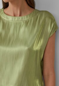 Grünes Satin-Oberteil mit rundem Ausschnitt und kurzen Ärmeln. Glatte Textur, leicht glänzend und mit subtilen Drapierungsdetails über dem Stoff.