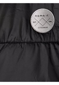 Černá prošívaná látka s prošívaným horizontálním švem a kulatou bílou nášivkou se značkou "name it EST. 1986 OUTERWEAR" v pravém horním rohu.