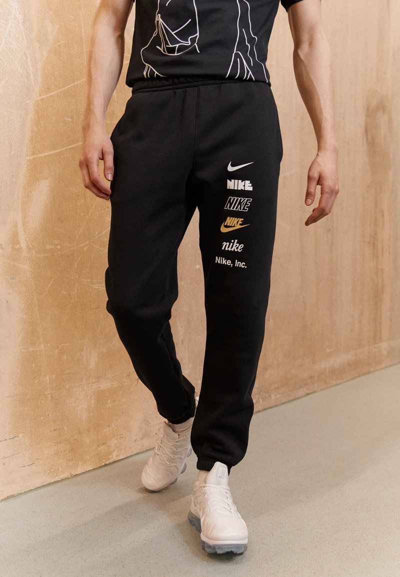 Pantalones deportivos negros con puños elásticos que presentan múltiples logotipos de Nike en blanco y amarillo. Llevados con zapatillas deportivas blancas, sobre un fondo claro.