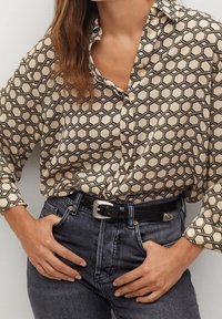Femme portant une chemise beige à motifs boutonnée, rentrée dans un jean taille haute foncé avec une ceinture noire à boucle argentée. Mains posées sur les hanches.