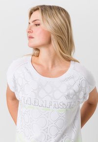 Witte korte mouwen top met een gestructureerd patroon van cirkels, voorzien van zilveren accenten en een logo in strass over de voorkant.