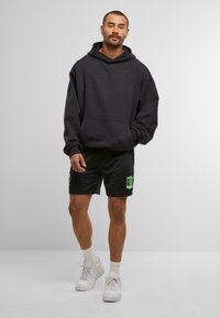 Mann, der einen schwarzen übergroßen Hoodie, schwarze Sport-Shorts mit einem grünen und gelben Emblem, weiße Socken und weiße Turnschuhe vor einem einfarbigen Hintergrund trägt.