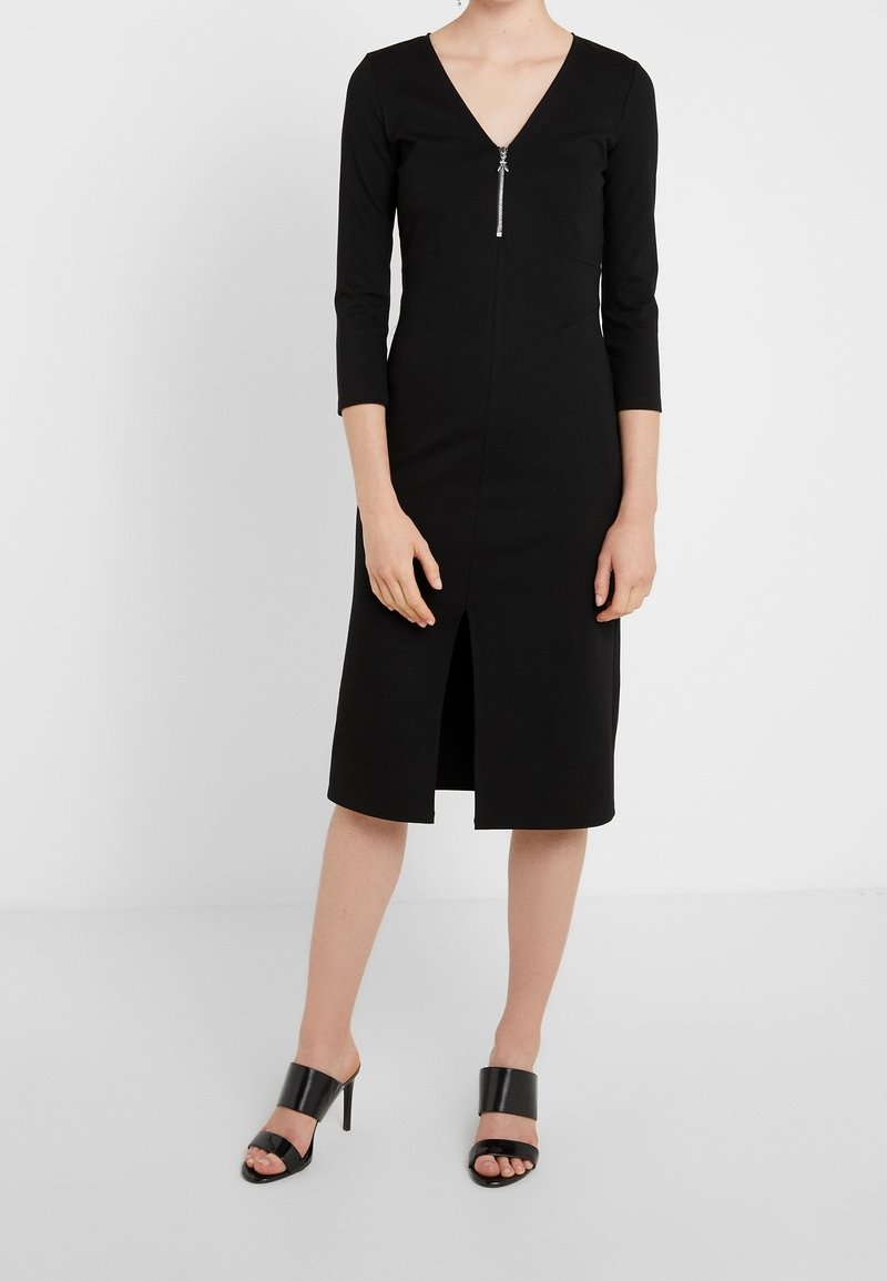Femme portant une robe noire jusqu'aux genoux avec des manches trois quarts, une fermeture éclair devant, une fente avant et des sandales noires à talons hauts.