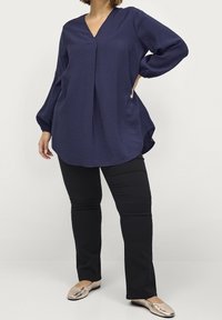 Blouse bleu marine avec un décolleté en V, à manches longues et en tissu texturé. Associée à un pantalon noir et des ballerines métalliques, avec une coupe décontractée.