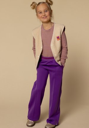 Witte fleece vest over een maroon en witte gestreepte longsleeve, gecombineerd met ruime paarse broek en zilveren schoenen.