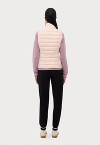 Kvinna med flätat hår, iförd en ljusrosa vadderad väst, lila långärmad tröja, svarta byxor och beige sneakers, som står med ryggen mot kameran.