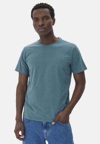 Camiseta de manga corta en color verde azulado, confeccionada en una suave mezcla de algodón, con un escote redondo y un diseño ajustado. Combinada con jeans de mezclilla azul.