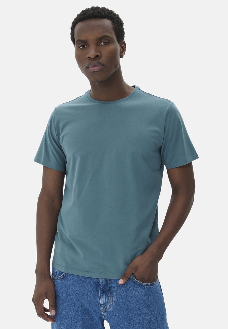 Camiseta de manga corta en color verde azulado, confeccionada en una suave mezcla de algodón, con un escote redondo y un diseño ajustado. Combinada con jeans de mezclilla azul.