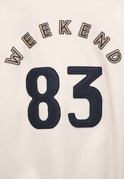 Μπεζ ύφασμα με κεντημένο κείμενο «WEEKEND» με καφέ περίγραμμα πάνω από μεγάλα μαύρα ψηφία «83».