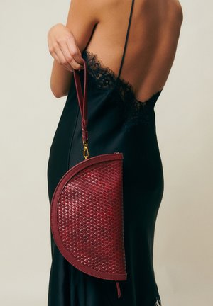 Frau in einem schwarzen Kleid mit Spitzenbesatz, die eine gewebte, halbmondförmige burgunderrote Clutch an ihrem Handgelenkriemen vor einem neutralen Hintergrund hält.