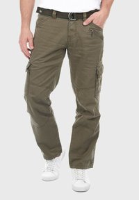 Olivgrüne Cargo-Hosen mit mehreren Taschen, strukturiertem Stoff und verstärkten Nähten. Kombiniert mit einem schwarzen Gürtel und weißen Sneakern.