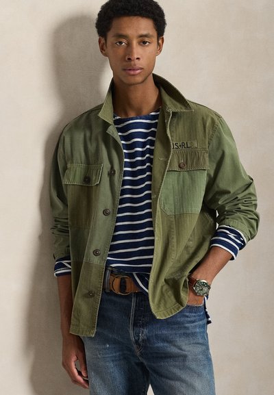 Πράσινο μπουφάν από καμβά με κουμπιά μπροστά και τσέπες στο στήθος, φορεμένο πάνω από μια μπλούζα με μπλε-ναυτικό και λευκές ρίγες. Συνδυάζεται με μπλε τζιν και καφέ ζώνη.