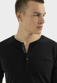 Schwarzes Langarm-Henley-Shirt mit Knopfleiste, aus strukturiertem Stoff und mit kleinem Logo auf der Brust.