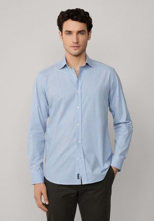 Camisa de manga larga a rayas en azul claro con cuello abotonado, que presenta una textura suave y una etiqueta en el dobladillo. Combinada con pantalones oscuros.