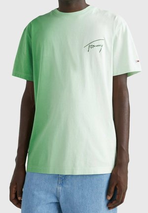 Homme portant un t-shirt vert clair Tommy Hilfiger avec logo sur la poitrine et la manche, associé à un jean en denim bleu.