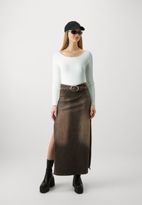 Fustă lungă din denim maro, cu o fente laterală și cataramă circulară, asociată cu un top alb cu mâneci lungi și accesorii negre.