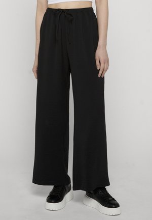 Person iført høytlivs svarte vide bukser med snor og svarte plattformsneakers på en ensfarget lys bakgrunn.