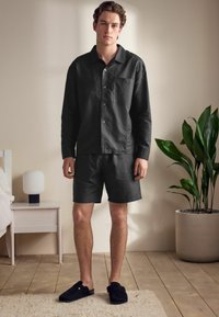 Black linen long-sleeve shirt with a pocket, paired with black shorts. Model wears dark slip-on shoes, standing on wooden floor.