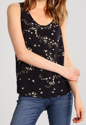 Femme portant un haut noir sans manches avec des motifs floraux et d'oiseaux blancs, associé à un jean bleu, bras croisés à la taille.