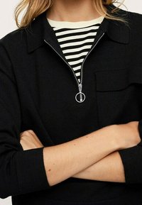 Personne portant un haut noir zippé par-dessus une chemise rayée noir et blanc, bras croisés au niveau de la taille, fermeture éclair partiellement ouverte.