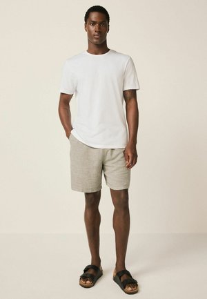 Mann steht und trägt ein schlichtes weißes T-Shirt, beige Shorts und schwarze Sandalen mit Schnalle vor neutralem Hintergrund.
