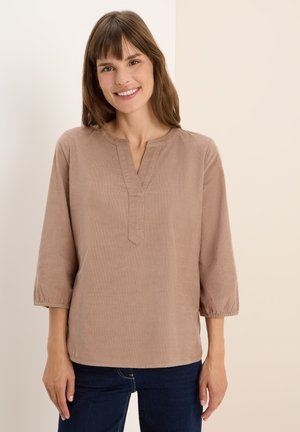 Jeune femme souriante, portant un haut à manches longues beige avec un décolleté en V et un jean bleu foncé, se tenant devant un fond clair.