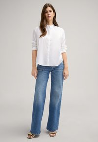 Weißes Hemd mit Stehkragen, kombiniert mit hellblauen, weiten Jeans und tanfarbenen, gehöhten Sandalen. Glattes Material, lässige Passform.