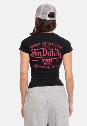 Personne portant un t-shirt noir ajusté Von Dutch avec un texte de logo rose, un pantalon de survêtement gris et une casquette de baseball en mesh gris clair, vue de dos.