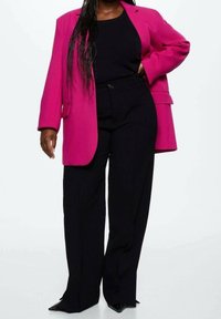 Helles pinkes Oversized-Blazer mit langen Ärmeln, kombiniert mit schwarzen hochtaillierten Hosen und spitzen schwarzen Absätzen. Glatte Stoffstruktur.