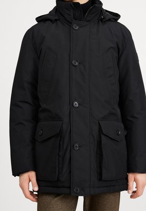 Homme portant une parka d'hiver noire boutonnée avec capuche et poches à rabat, debout devant un fond clair uni.
