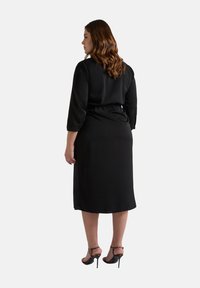 Robe noire jusqu'aux genoux avec des manches trois-quarts, taille élastique pour un ajustement réglable et tissu doux. Portée avec des talons hauts à lanières.