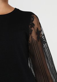 Top nero con scollo rotondo e maniche lunghe in pizzo trasparente con motivi floreali. Il corpo è realizzato in un tessuto solido a coste.