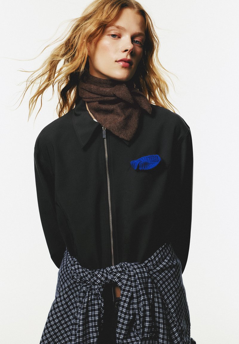 Une jeune femme aux cheveux roux ondulés porte une veste à zip noire, une écharpe marron et une chemise à carreaux bleue nouée autour de la taille, contre un fond blanc.