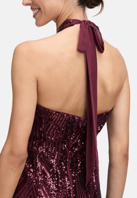 Frau in einem rückenfreien, gepaarten Halterneck-Kleid in Dunkelviolett, mit einer Schleife am Hals gebunden.