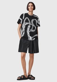 Svart t-shirt med vitt abstrakt mönster, kombinerad med svarta lädershorts. Modellen bär svarta sandaler och ett silverfärgat kedjehalsband.