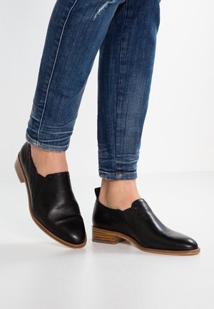 Sorte lædersko uden snørebånd med lave træhæle, båret med blå skinny jeans på en hvid gulvbaggrund.